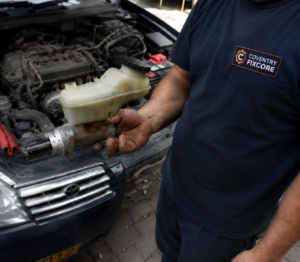 Close-up automotive repair scene showing a mechanic in Coventry Fixcore uniform holding a brake master cylinder while inspecting a car engine in a professional workshop environment.